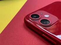 A close-up view of a red smartphone focusing on the dual camera lenses. The phone is placed on a red surface, with a contrasting yellow background. The shiny surface of the phone reflects light, emphasizing its glossy finish.